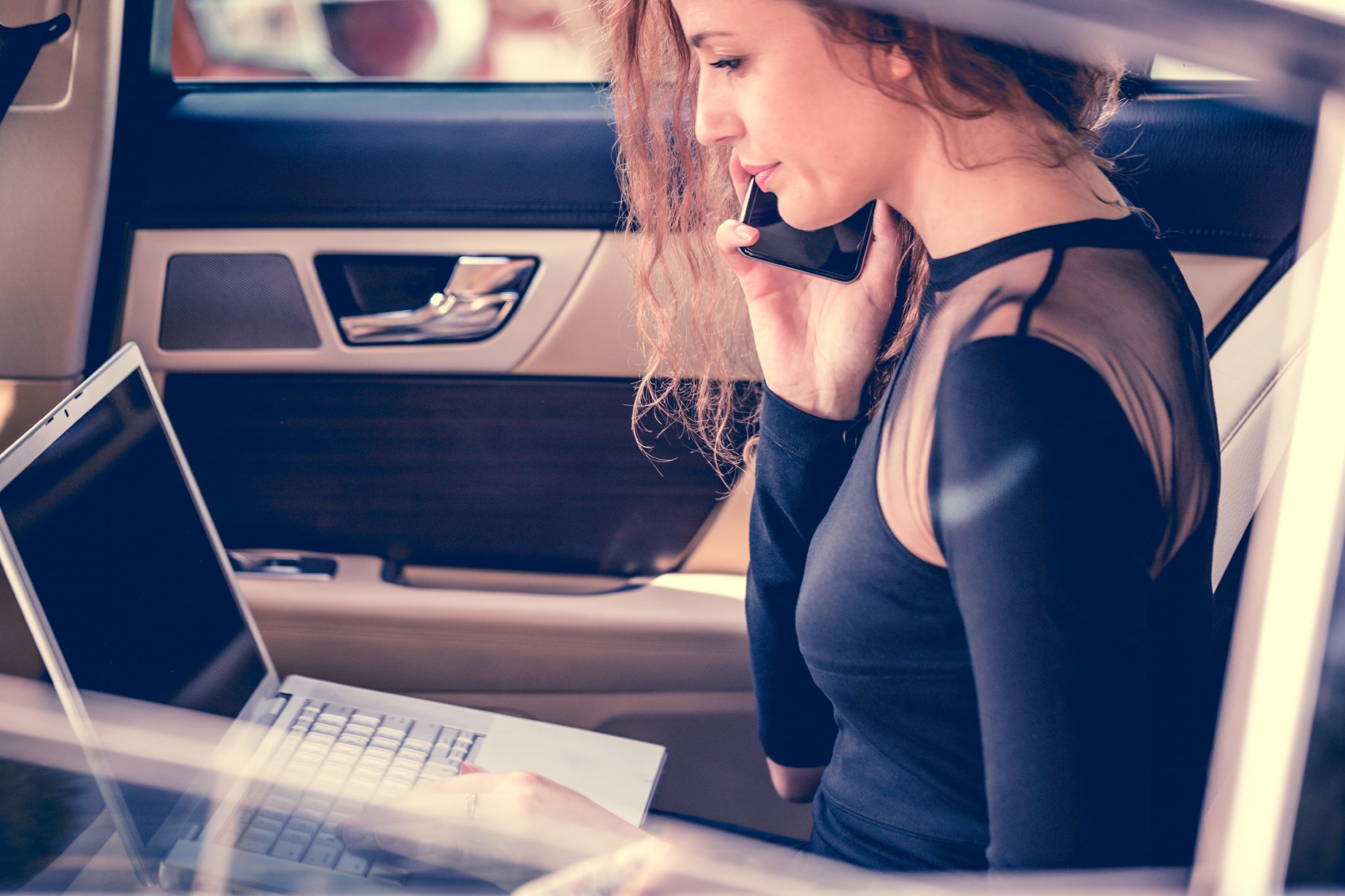 European girl in her car multi-tasking, using her laptop while talking on phone.