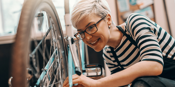 Beautiful Caucasian female worker with short blonde hair and eyeglasses crouching and repairing bicycle. Bike workshop interior.