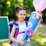 Happy,Little,Kid,Girl,By,Desk,With,Backpack,Or,Satchel