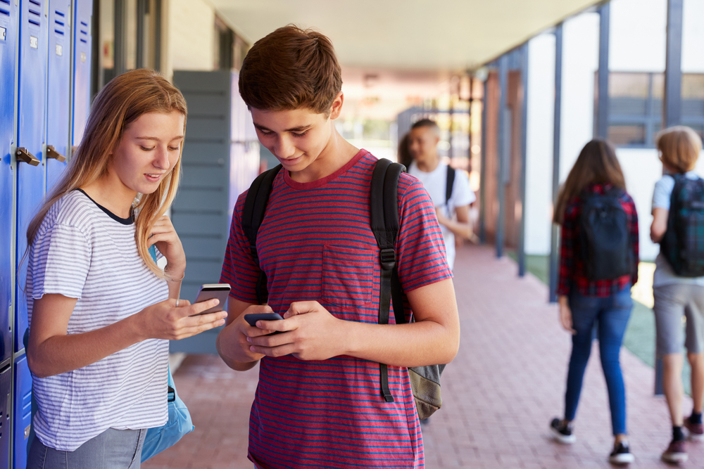 Two,Friends,Talking,And,Using,Phones,In,School,Corridor