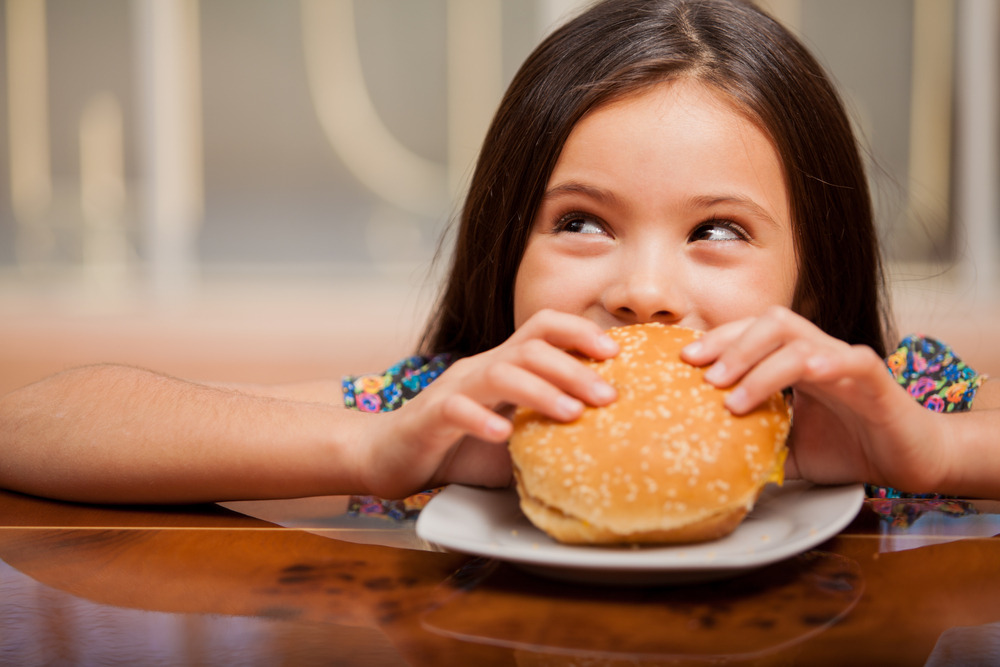 Cute,Little,Latin,Girl,Eating,A,Hamburger,And,Looking,Up