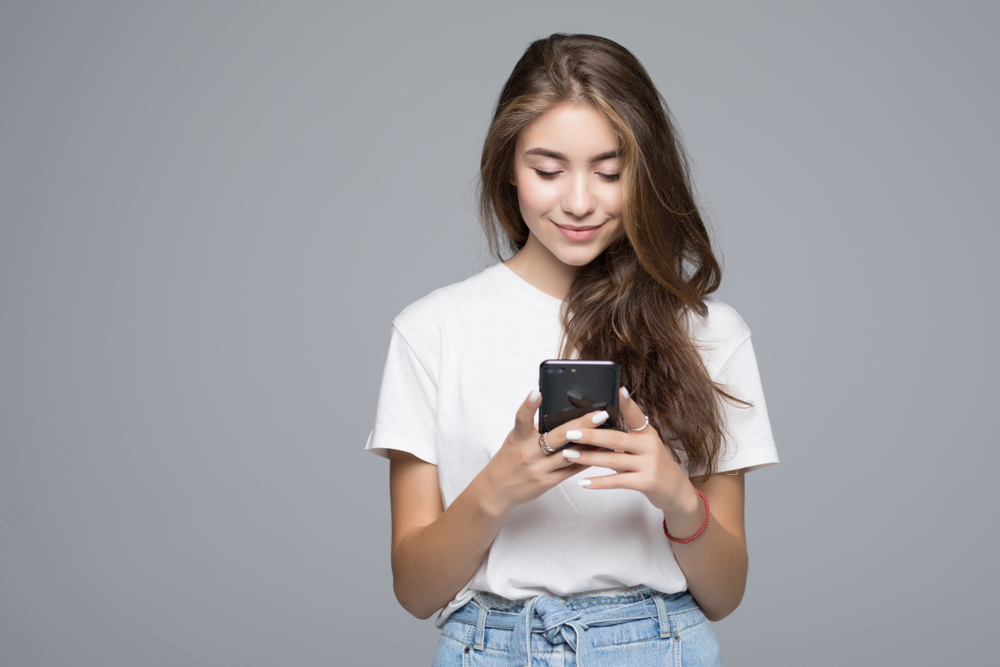 Smiling,Lovely,Young,Woman,Standing,And,Using,Cell,Phone,Over